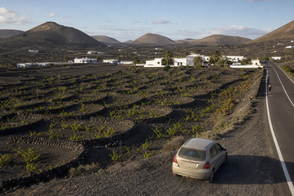 Road winding through black volcanic vineyards with stone walls and a single car