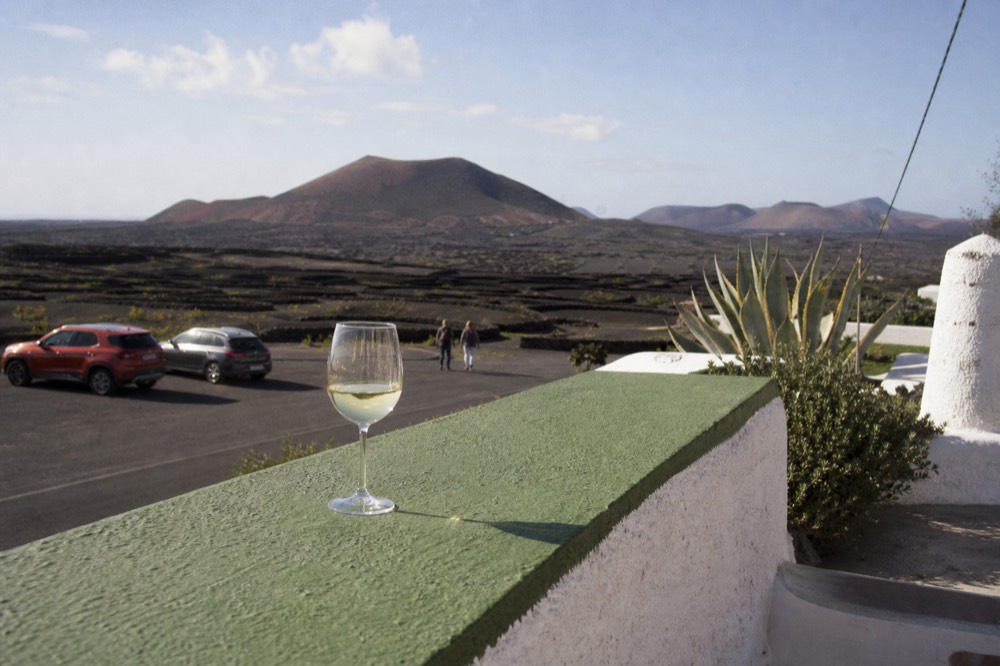 A glass of white wine on a sunlit terrace overlooking black vineyard ash fields