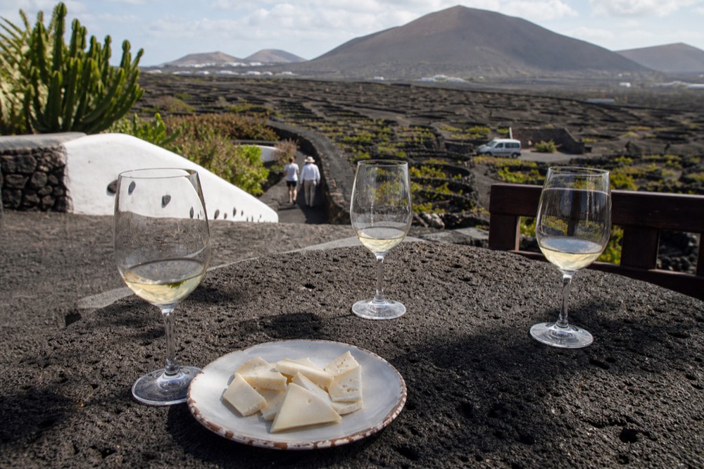 A tasting flight of white wines and a plate of cheese on a bodega terrace overlooking volcanic cones