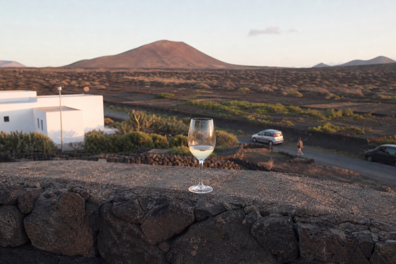 A glass of white wine resting on a stone wall above black volcanic ash vineyards