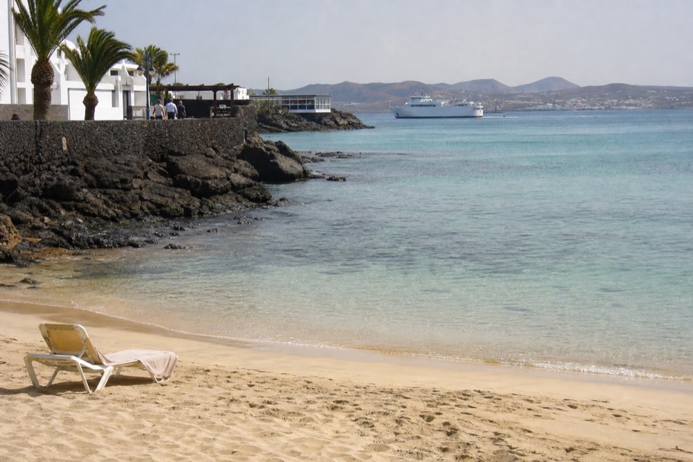 A calm sandy beach in Playa Blanca with clear shallow water and a distant ferry