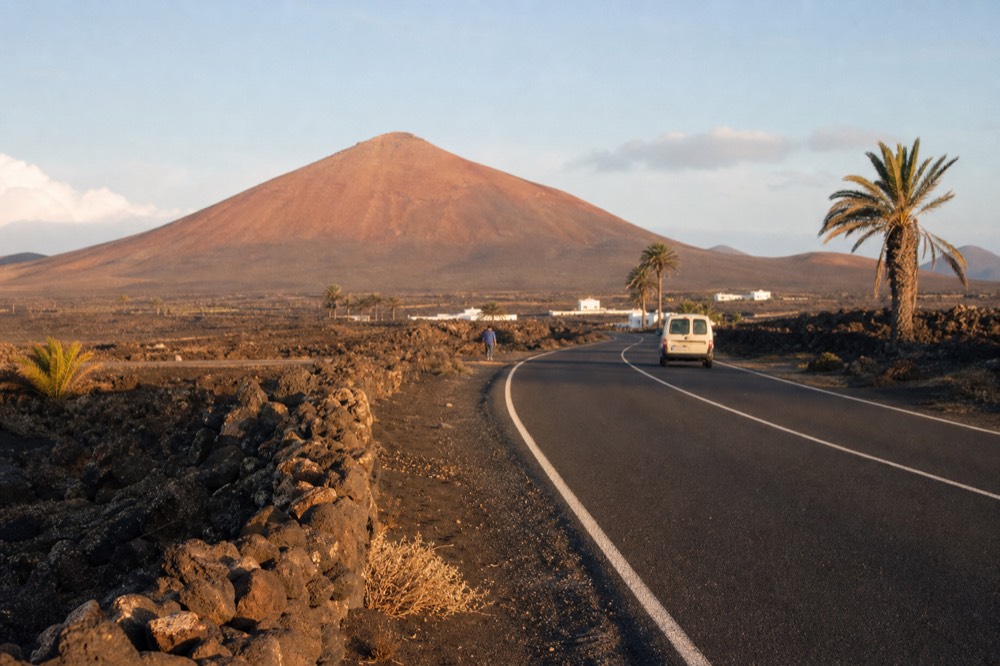 A quiet two-lane island road curving through black ash fields with a low volcano on the horizon