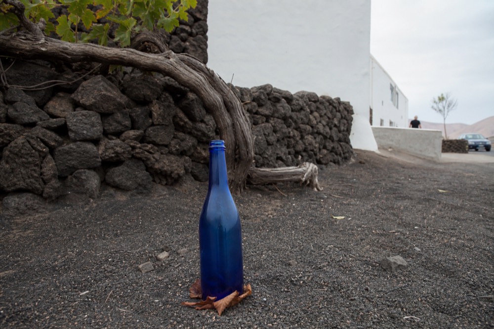 Blue glass wine bottle standing in black volcanic sand with a vine pit behind