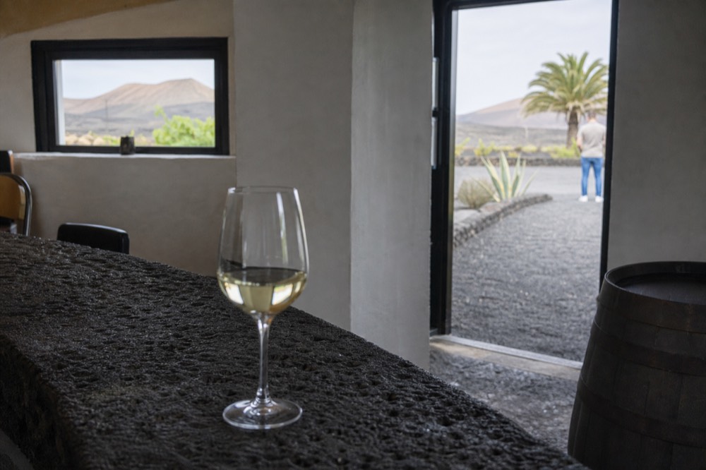 A glass of pale white wine on a stone counter with dark volcanic soil behind