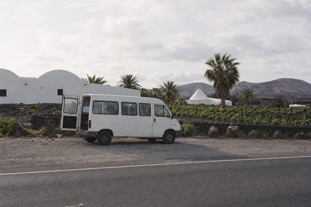A small minibus parked on a road beside black ash vineyards and a white winery building